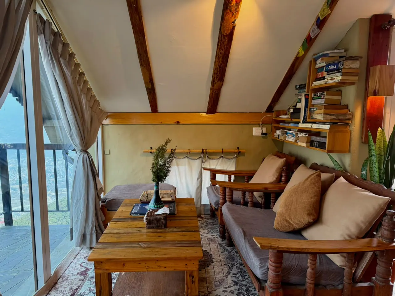 Sofa lounge in the Attic room with cushions, a wooden table and a bookshelf under timber roof beams, next to glass doors onto the veranda